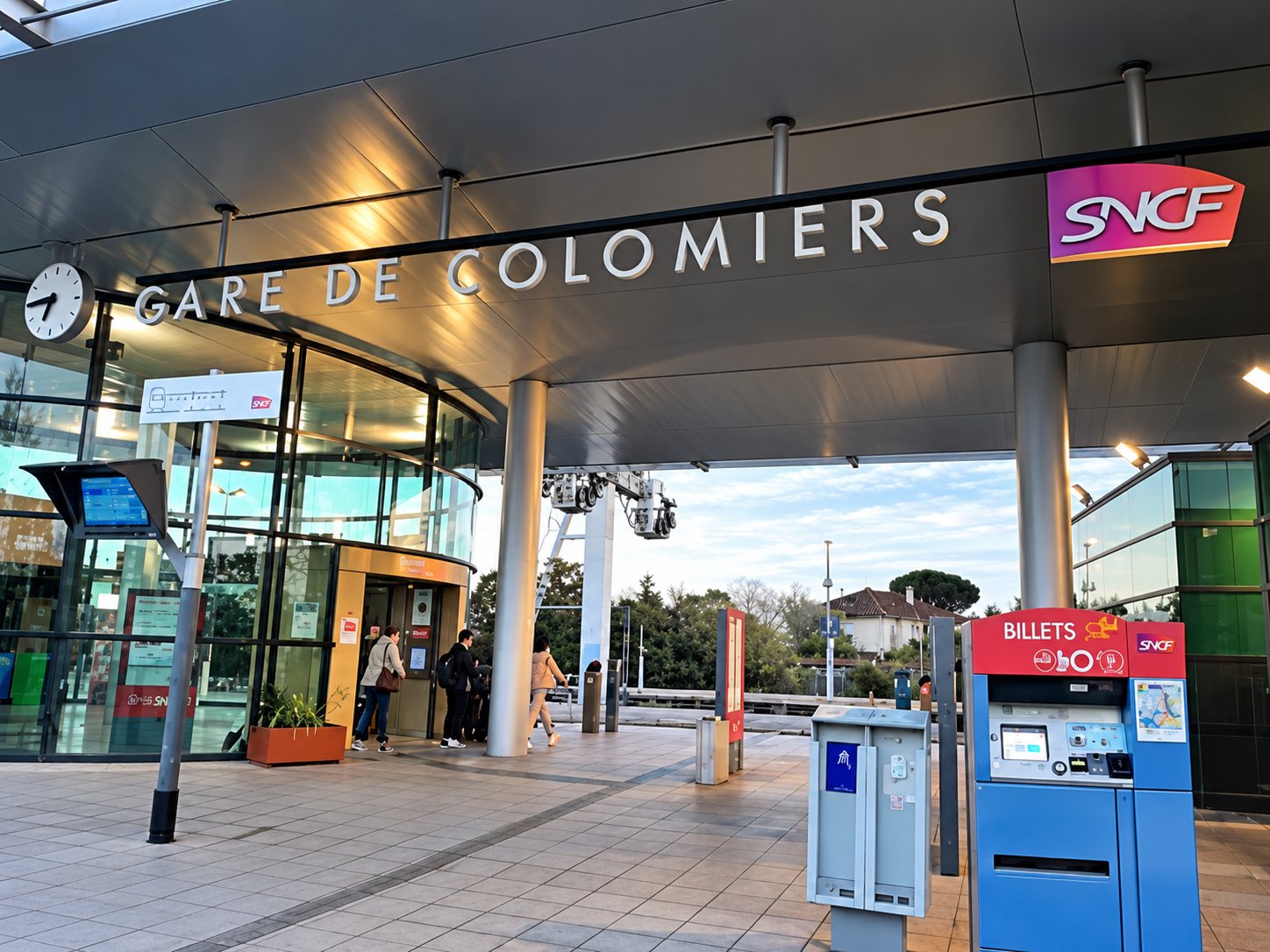 Façade de la gare SNCF de Colomiers sous un auvent, avec une borne bleue et rouge au premier plan à droite.